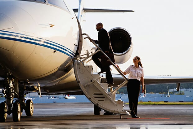 A pilot assists a passenger boarding a private jet on the tarmac, with the aircraft's sleek design and engines visible.