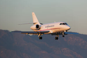 A sleek private jet descends against a backdrop of mountains during sunset, showcasing its elegant design and landing gear.