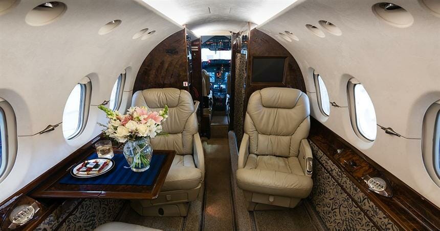 Luxurious private jet interior featuring leather seats, a wooden table with flowers, and a cockpit visible in the background.