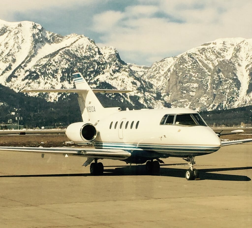 A private jet parked on a runway with snow-capped mountains in the background under a partly cloudy sky.
