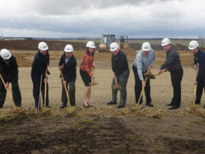 Group of people in hard hats using shovels for a groundbreaking ceremony on a construction site.