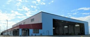 Large aviation hangar with a red and blue exterior, under a bright sky with scattered clouds.