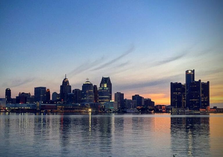 Detroit skyline at dusk, reflecting on calm waters under a colorful sunset sky.