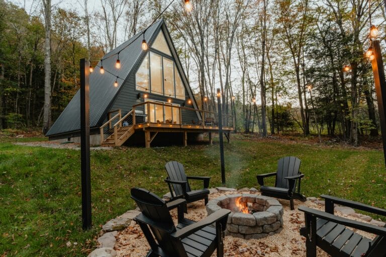 A modern A-frame cabin surrounded by trees, featuring a fire pit and string lights, creating a cozy outdoor atmosphere.