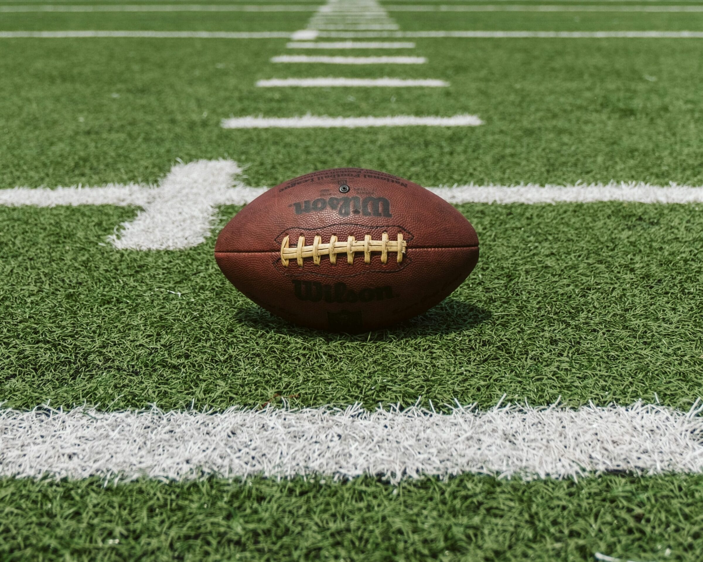 A brown football rests on green turf, with white yard lines visible in the background.