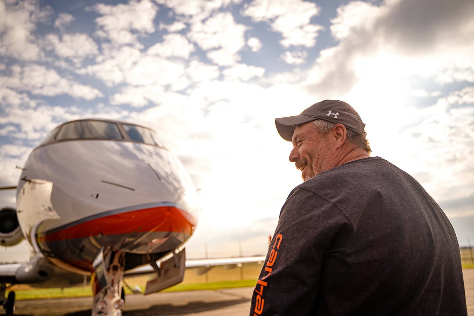 A smiling man in a cap stands beside a sleek jet, with a cloudy sky and sunlight creating a warm atmosphere.