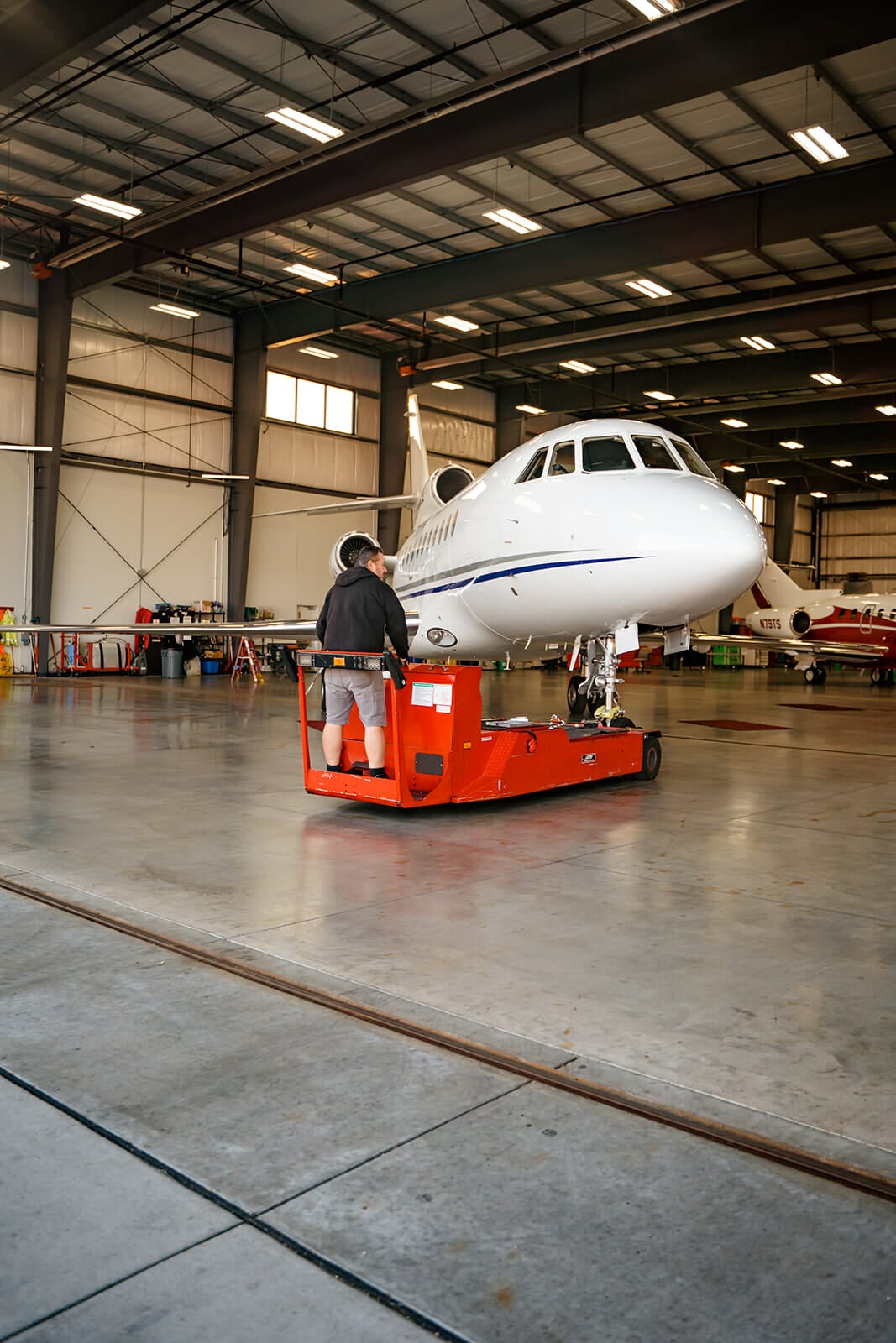 A technician operates a red maintenance cart near a sleek white jet in a spacious hangar with overhead lighting.