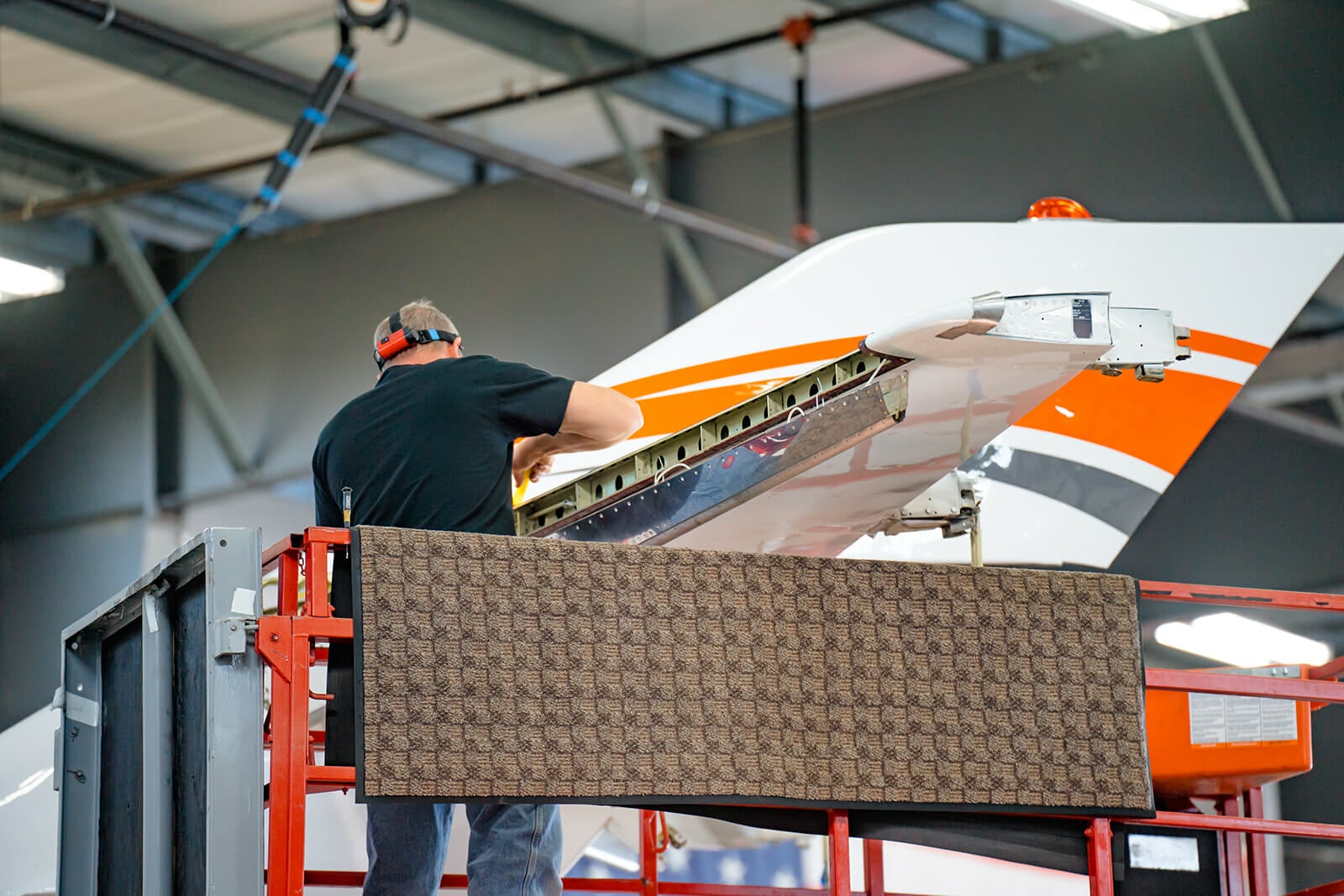 A technician works on the tail section of an aircraft while standing on a lift inside a hangar.