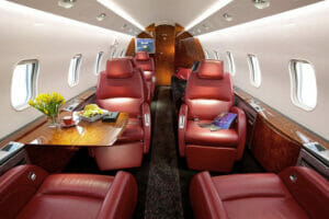 Luxurious private jet interior featuring red leather seats, a wooden table with flowers, and large windows.