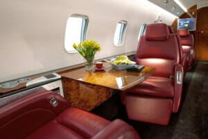 Luxurious private jet interior featuring red leather seats, a polished wooden table with flowers, and a gourmet meal.