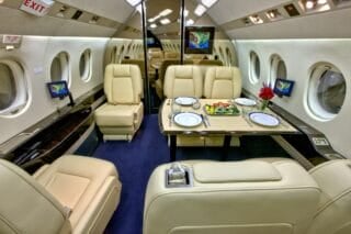 Luxurious private jet interior featuring beige leather seating, a dining table set with plates, and a floral centerpiece.
