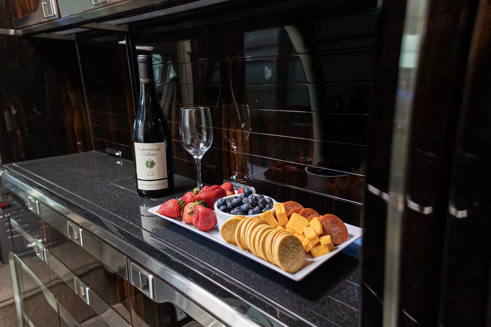 A bottle of Cakebread Cellars wine, a glass, and a platter of strawberries, blueberries, cheese, and crackers on a countertop.