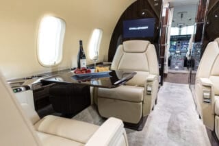 Luxurious private jet interior featuring cream leather seats, a polished table with wine, fruit, and a cockpit in the background.