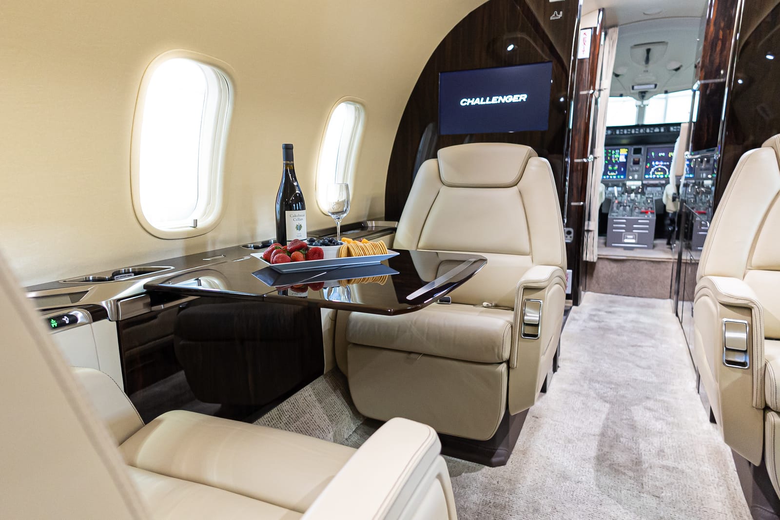 Luxurious private jet interior featuring cream leather seats, a polished table with wine, fruit, and a cockpit in the background.