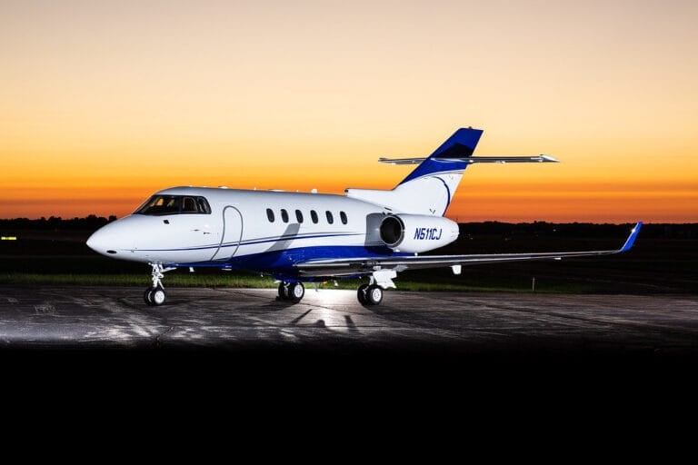 A sleek private jet with a blue and white design parked on an airstrip at sunset, casting long shadows.