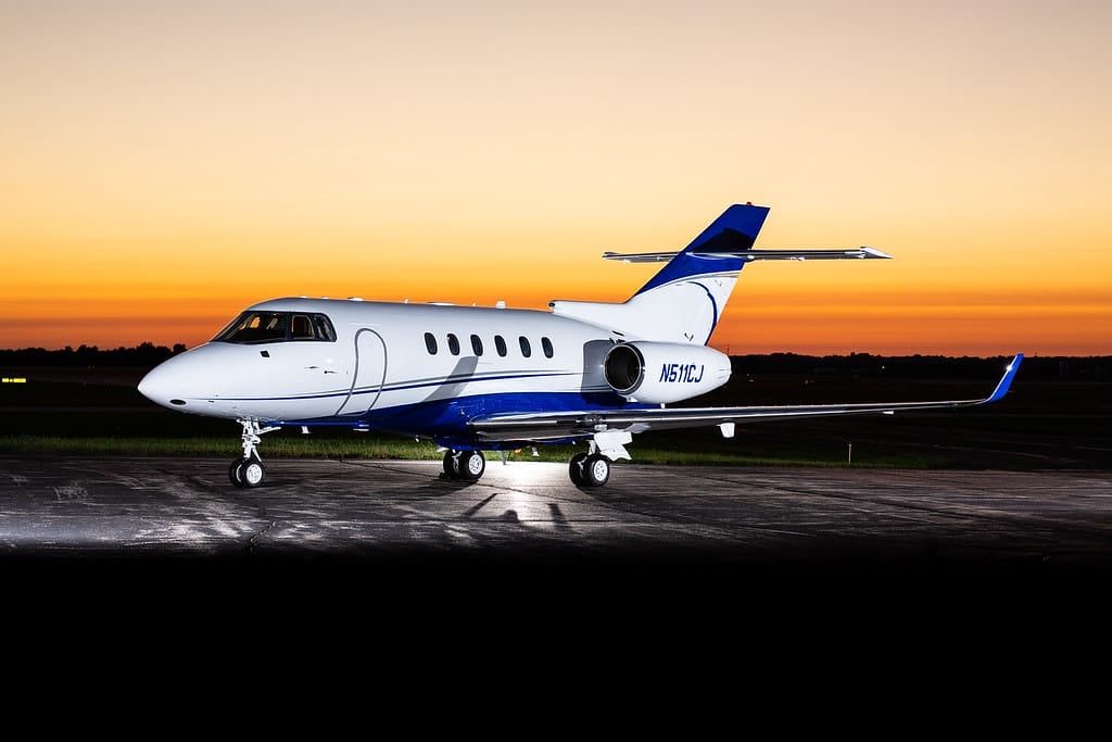 A sleek private jet with a blue and white design parked on an airstrip at sunset, casting long shadows.
