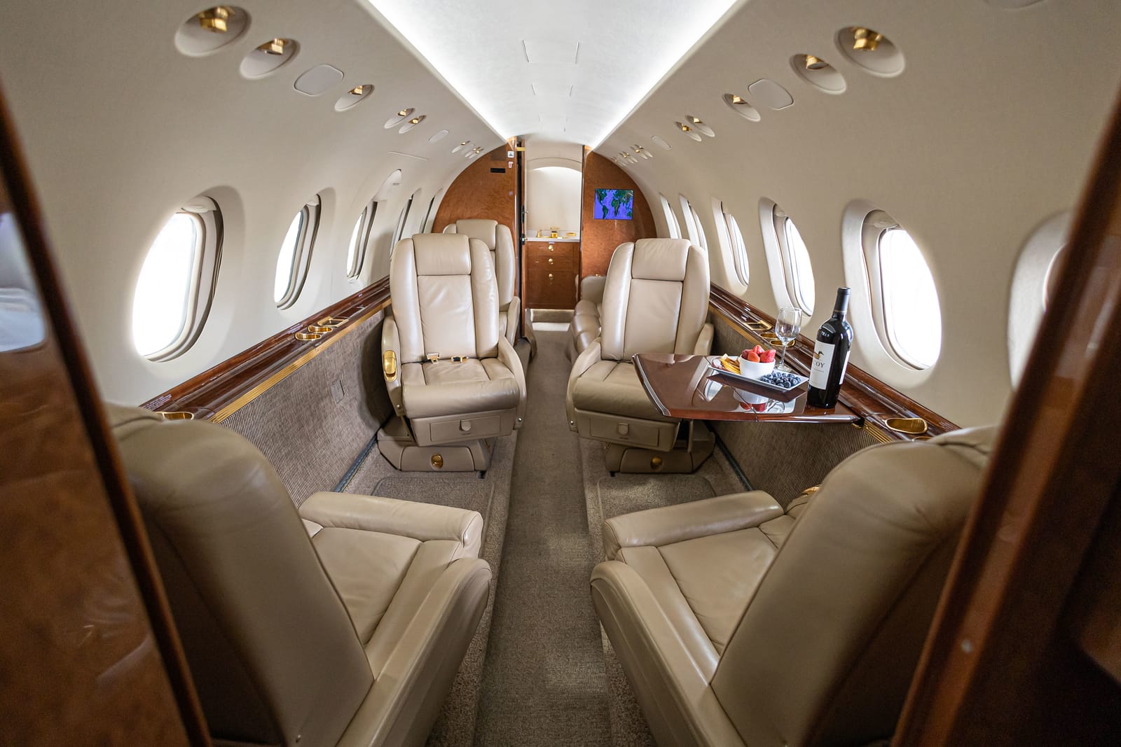 Luxurious private jet interior featuring beige leather seats, a wooden table with snacks, and large windows.