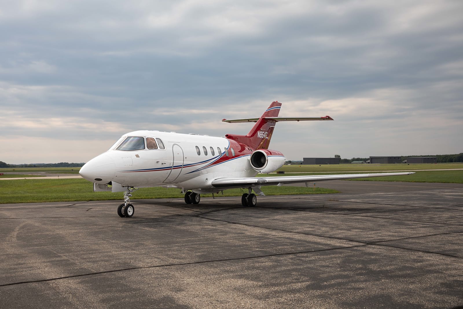 A sleek white and red private jet parked on a runway under a cloudy sky, surrounded by green grass and distant hangars.