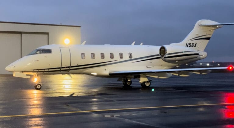 A sleek white private jet with black accents parked on a wet runway, illuminated by a nearby hangar light.
