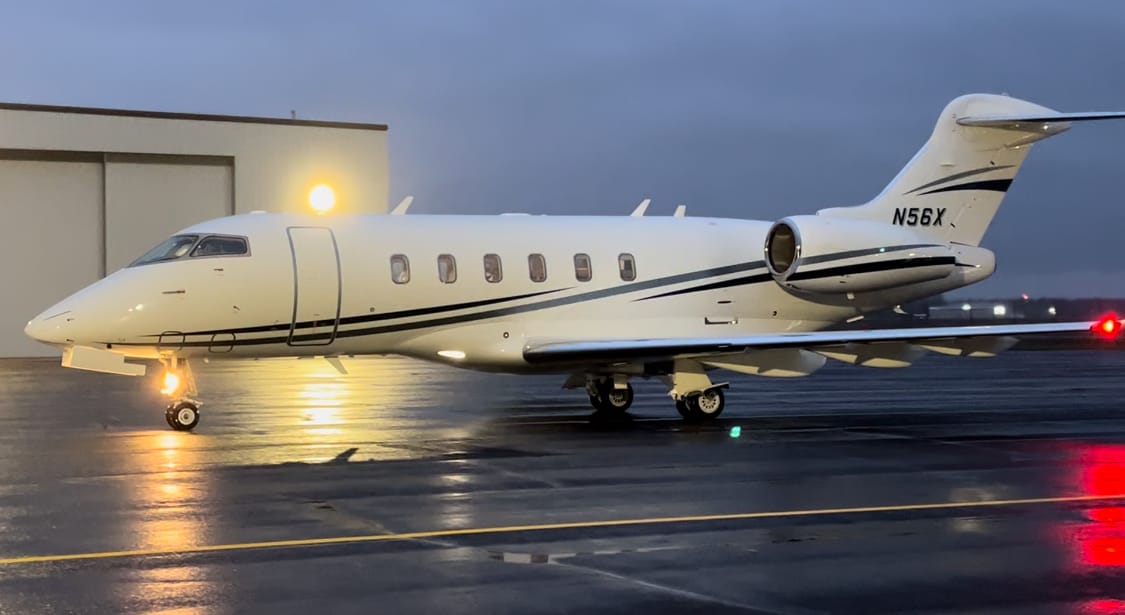A sleek white private jet with black accents parked on a wet runway, illuminated by a nearby hangar light.