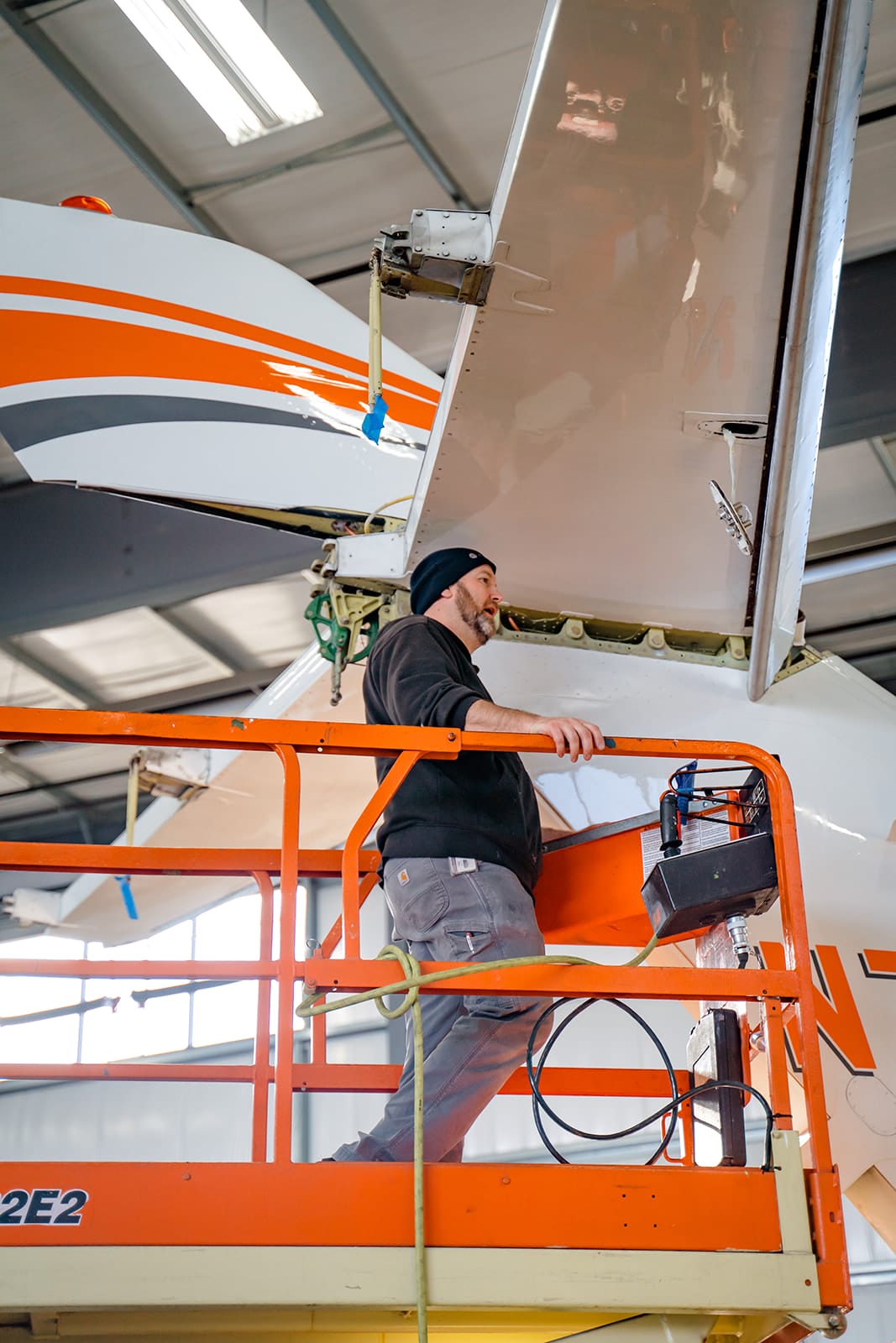 A technician on a lift inspects the tail of a helicopter with orange and gray accents in a hangar.