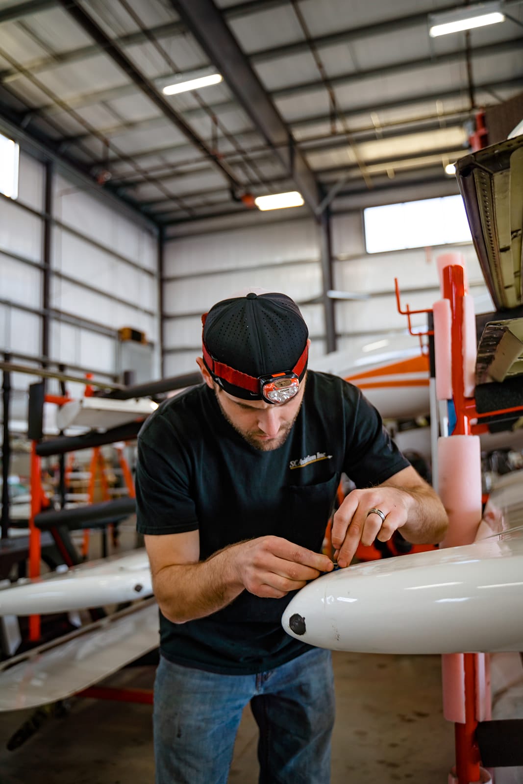 A man with a headlamp carefully works on a white aircraft part in a well-lit hangar filled with tools and equipment.