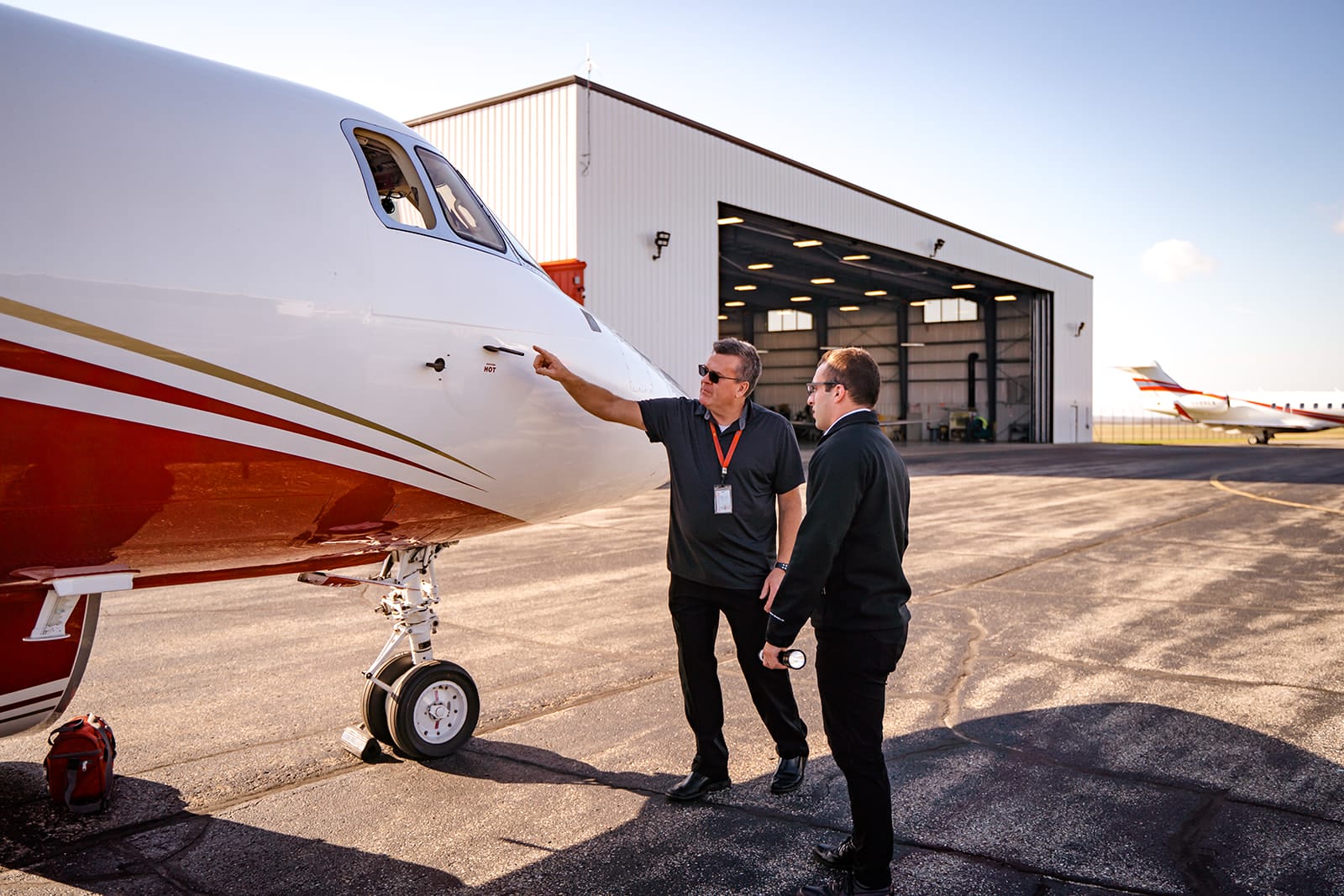 Two men inspect a private jet on the tarmac, discussing details near a hangar under a clear sky.
