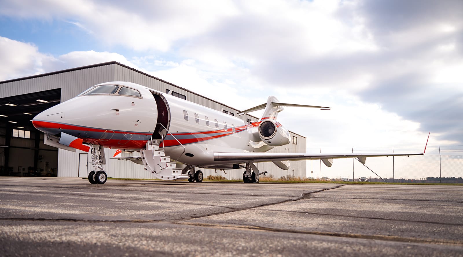 A sleek private jet with red accents parked on a tarmac beside a hangar under a cloudy sky.