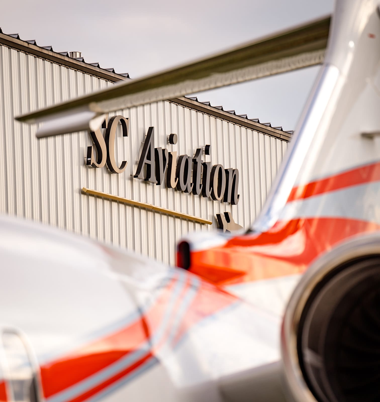 Tail of a silver and red aircraft in focus, with "SC Aviation" sign on a metal building in the background.