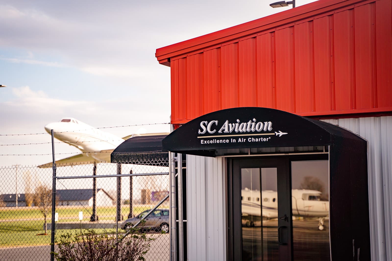 SC Aviation building with a black awning, featuring a white aircraft sculpture in the background against a cloudy sky.
