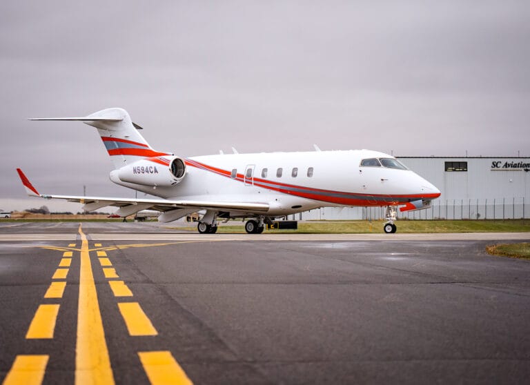 A sleek white private jet with red accents parked on a runway, with a hangar in the background under a cloudy sky.