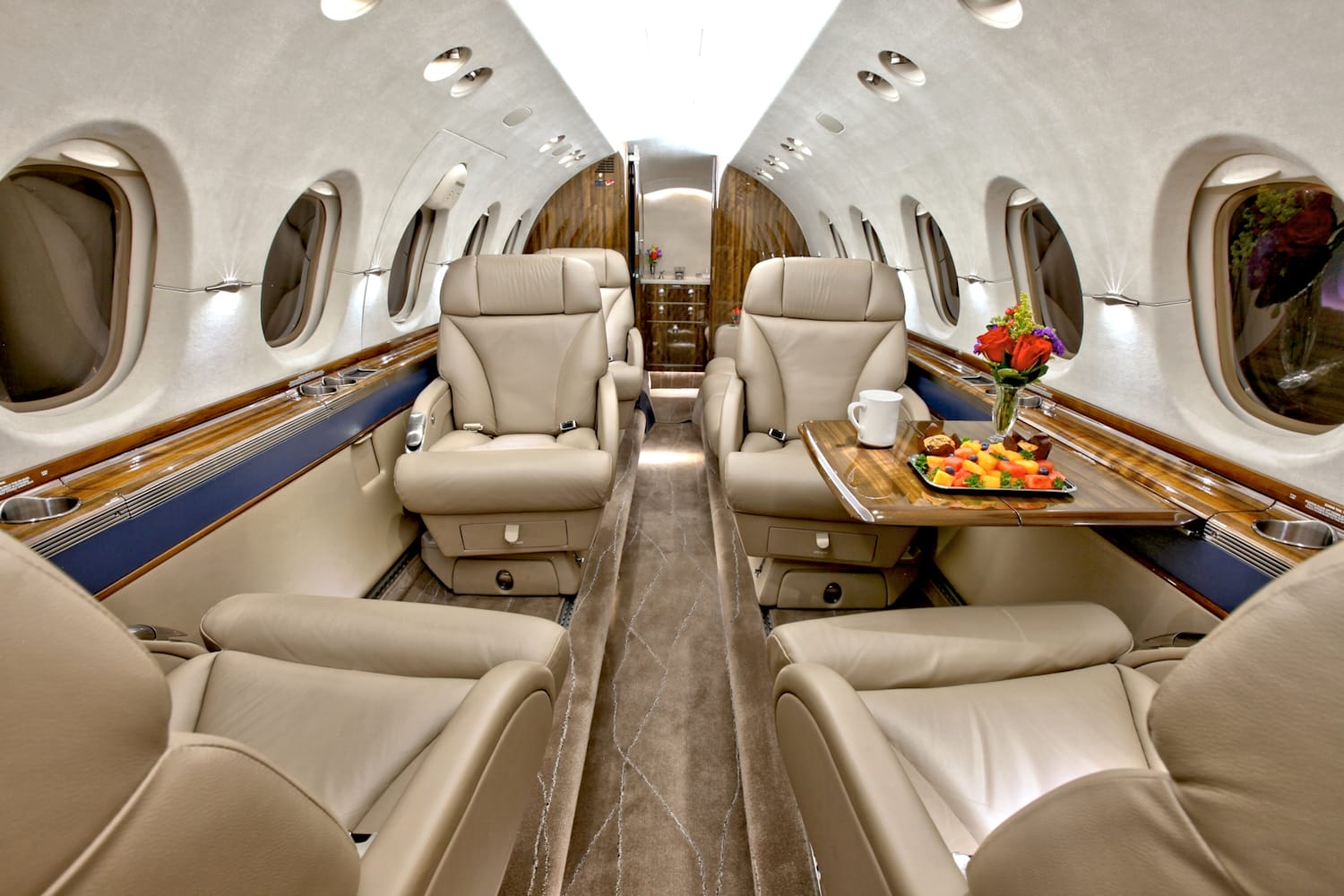 Luxurious private jet interior featuring plush beige seats, a wooden table with fruit and flowers, and large windows.