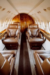 Luxurious private jet interior featuring plush brown leather seats and wooden accents along a narrow aisle.