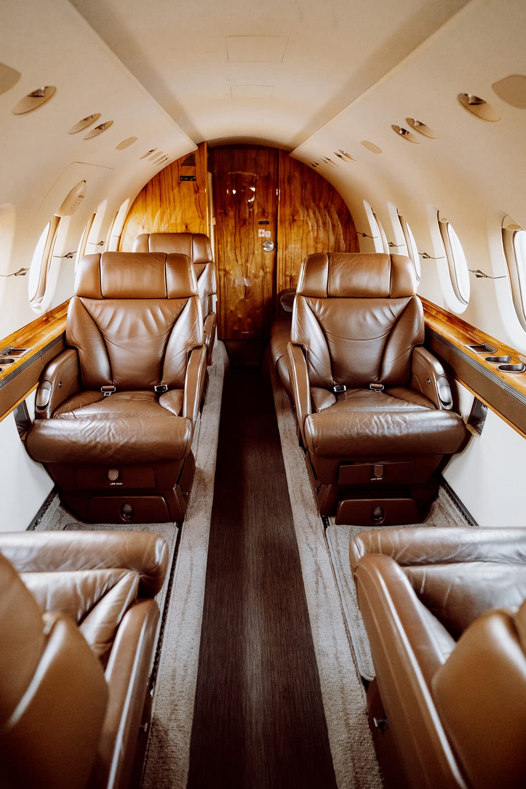 Luxurious private jet interior featuring plush brown leather seats and wooden accents along a narrow aisle.