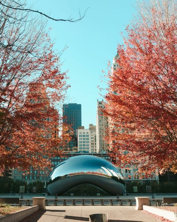 Reflective sculpture surrounded by vibrant autumn trees and a skyline of tall buildings under a clear blue sky.