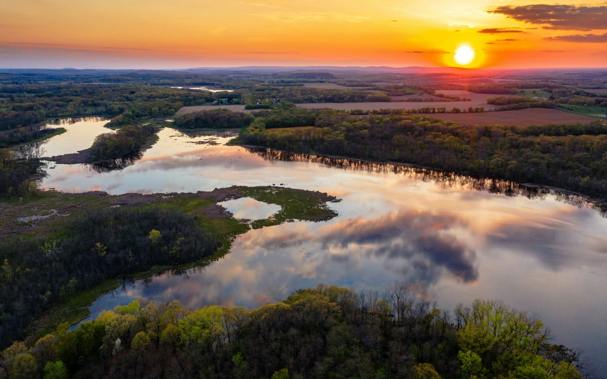 Sunset over a winding river, reflecting vibrant colors, surrounded by lush greenery and rolling hills in the background.