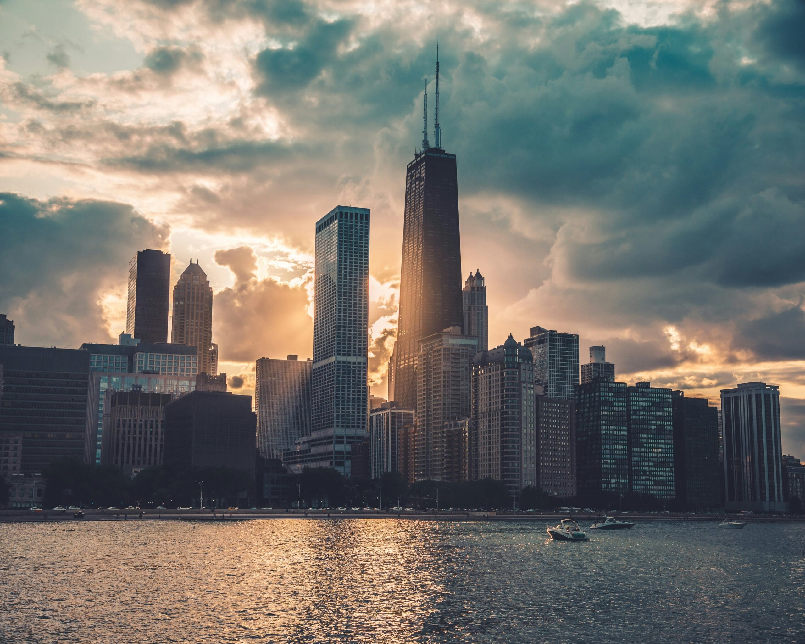 Chicago skyline at sunset, featuring tall skyscrapers silhouetted against dramatic clouds and shimmering water.