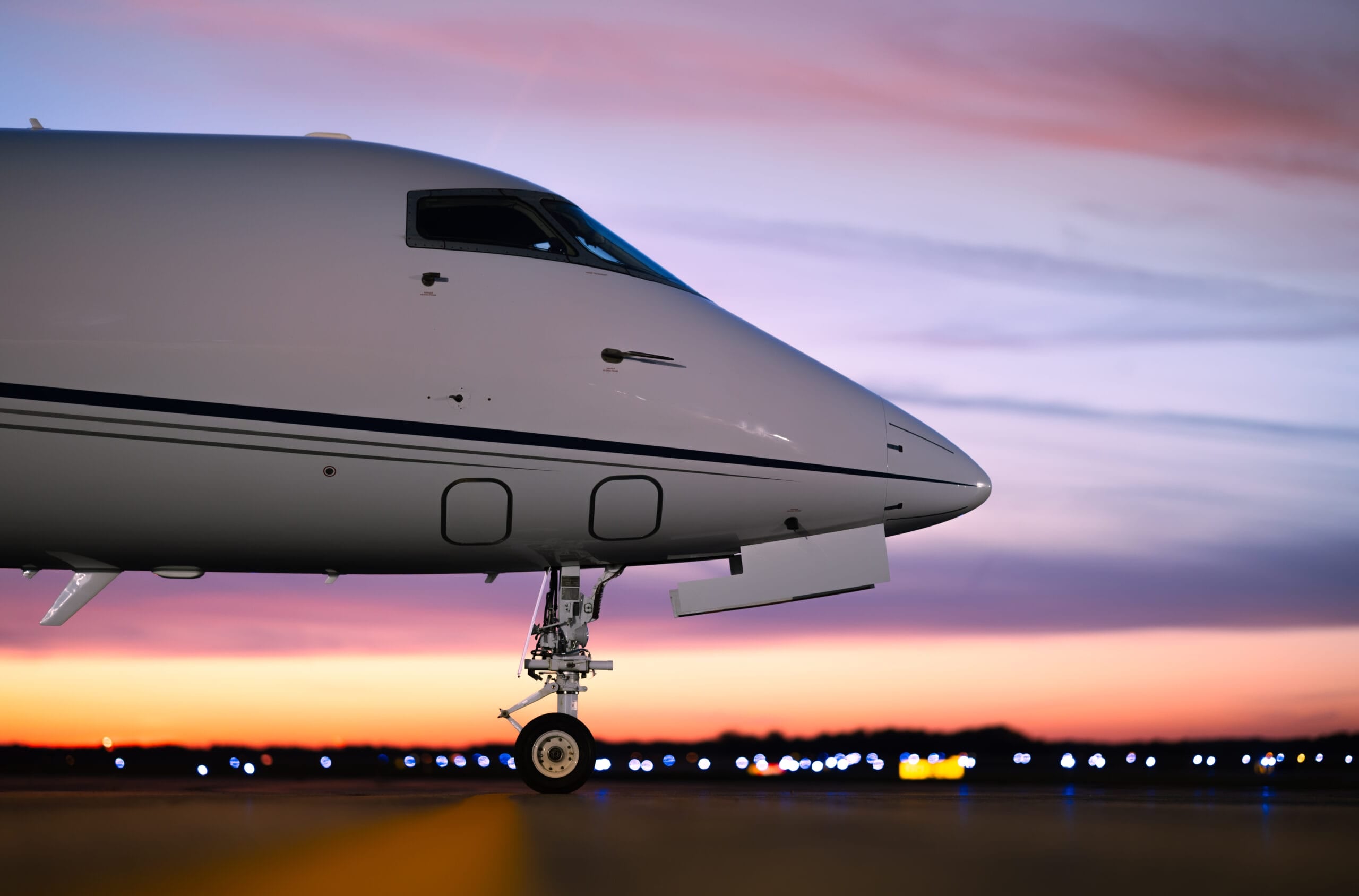 Nose of a sleek private jet against a vibrant sunset sky, with blurred runway lights in the background.