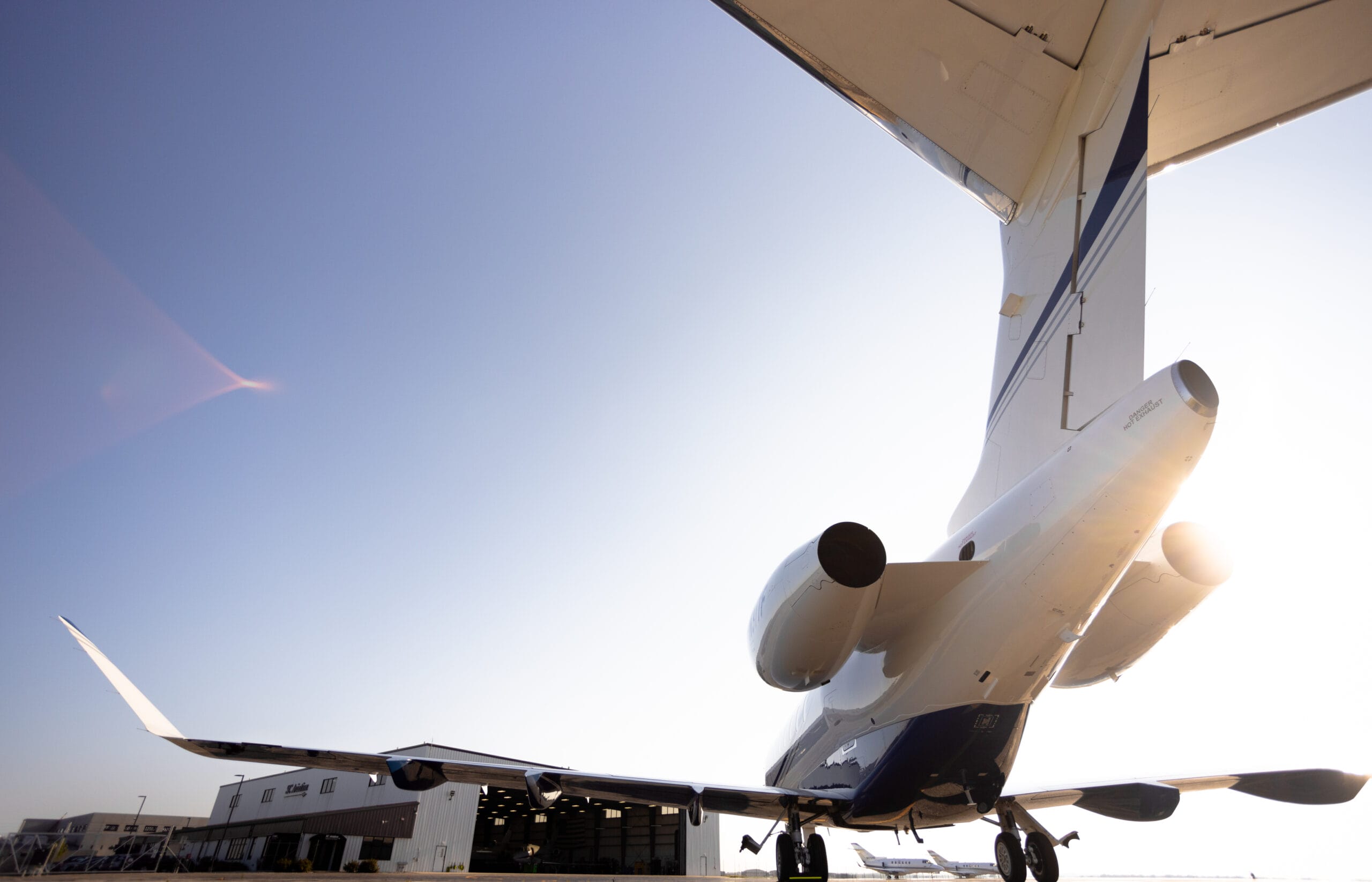 Business jet with prominent engines and tail, set against a clear blue sky and hangar in the background.