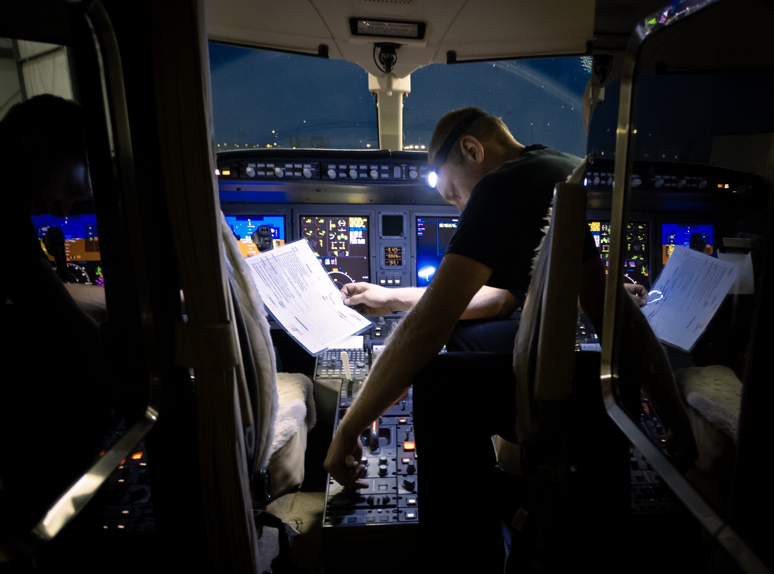Pilot reviewing documents in a dimly lit cockpit, surrounded by control panels and illuminated displays.