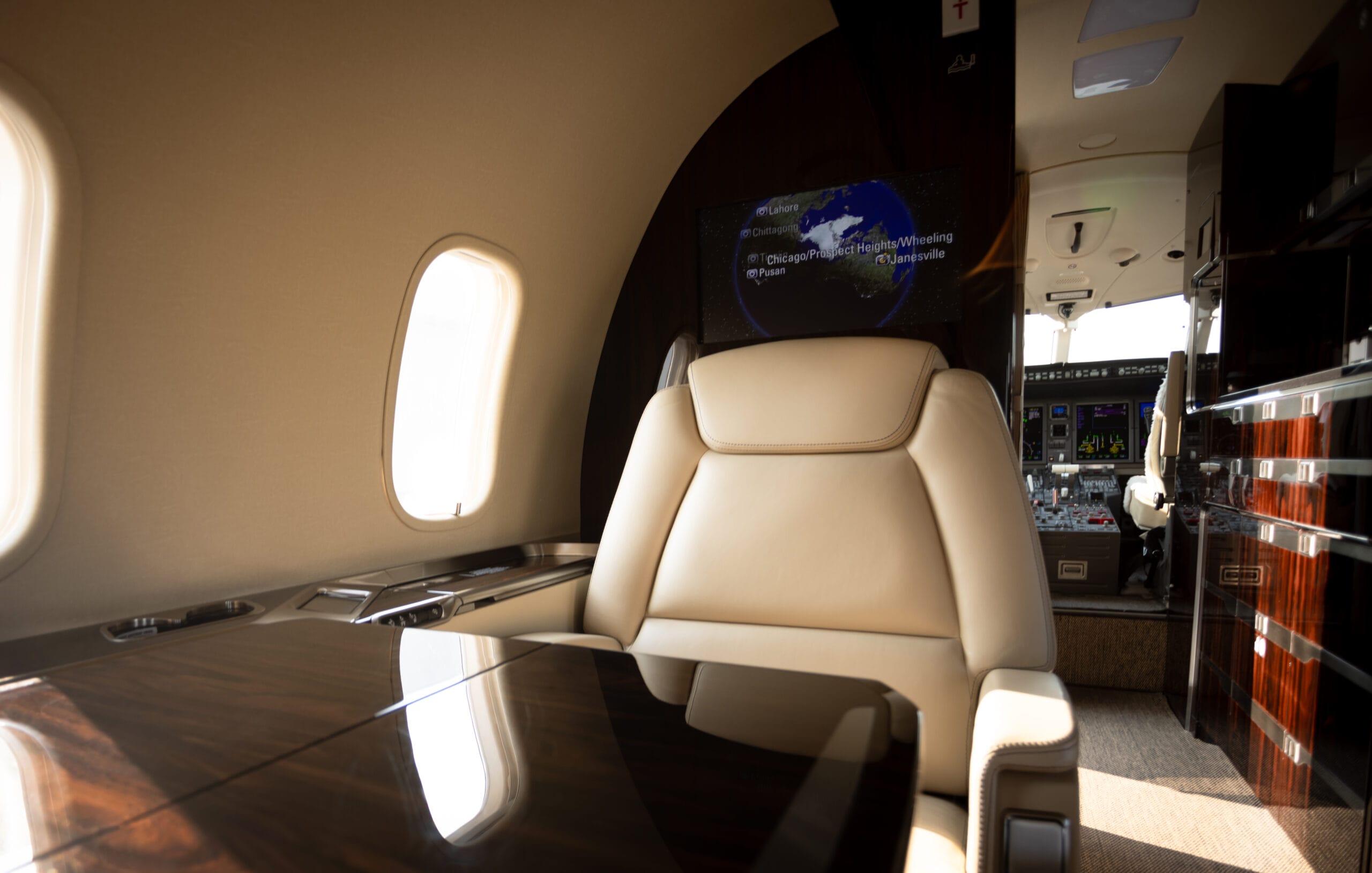 Luxurious private jet interior featuring a plush leather seat, polished wood table, and a cockpit visible in the background.