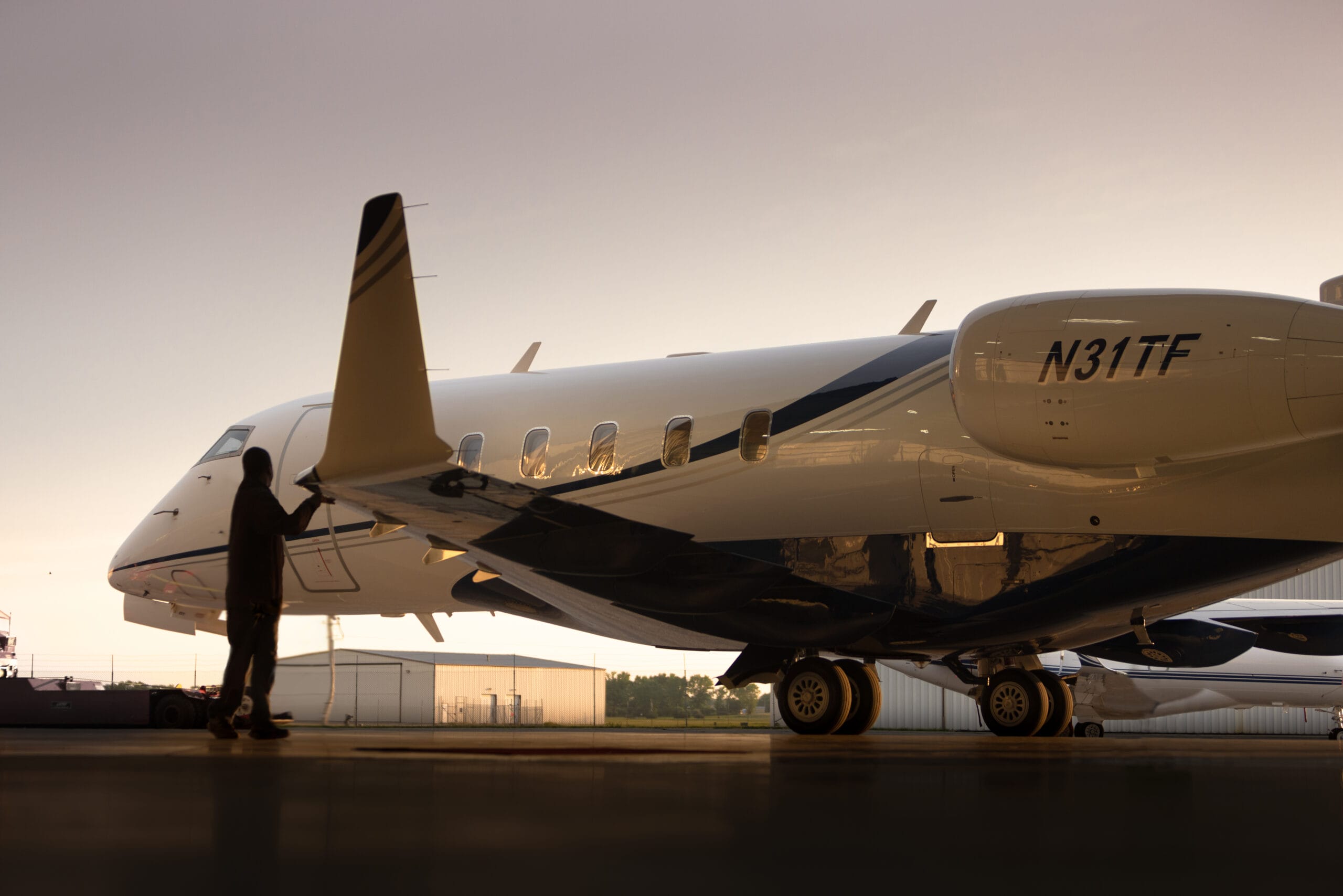 A silhouette of a person standing beside a sleek private jet in a hangar, illuminated by soft evening light.
