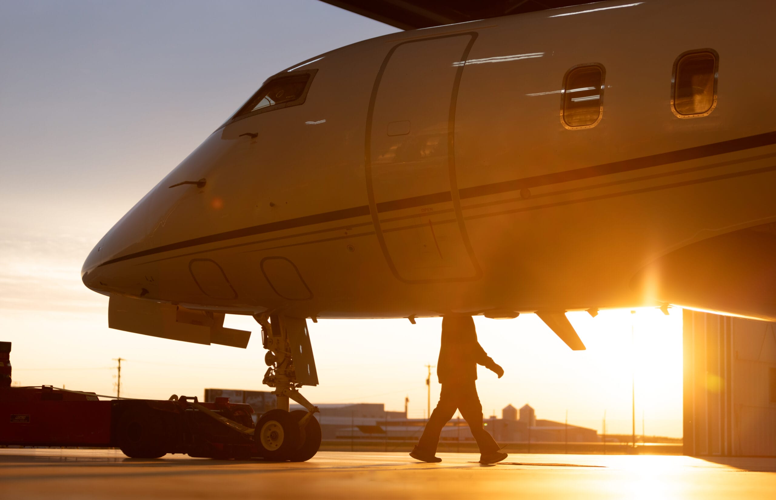 A silhouette of a person walking beneath a private jet, illuminated by a warm sunrise glow in a hangar.