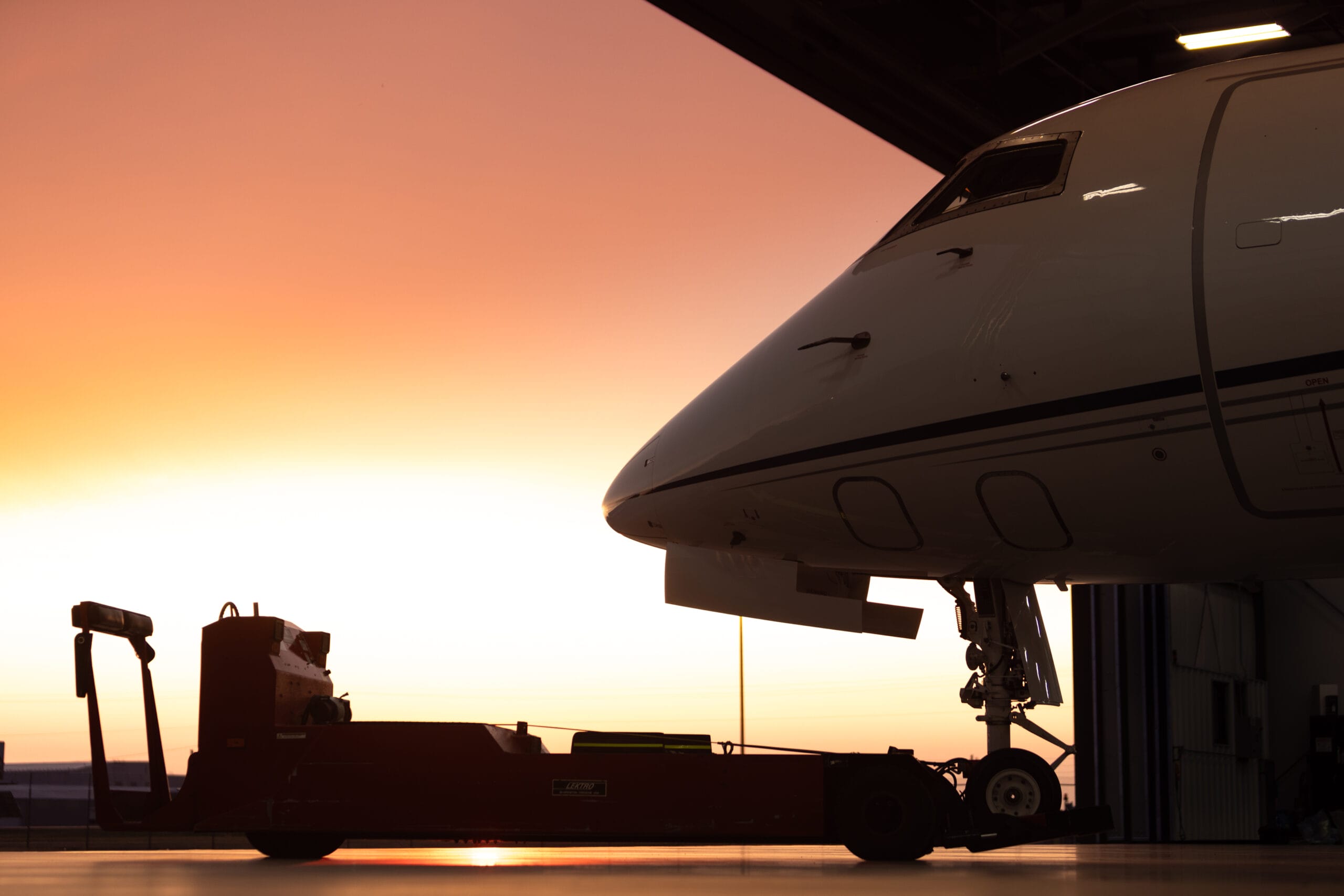 Silhouette of a jet aircraft in a hangar, with a red tow vehicle and a vibrant sunset in the background.