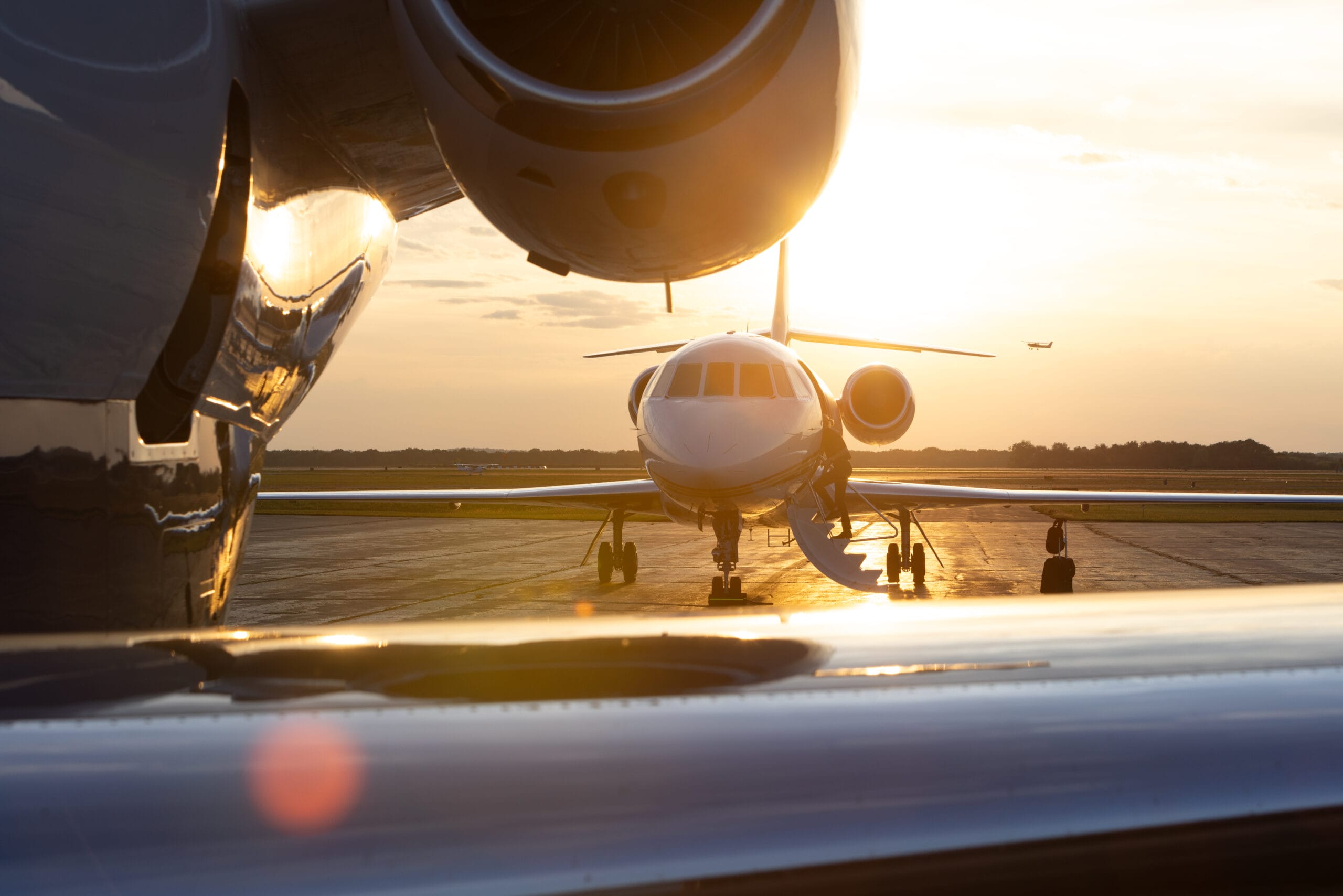 Private jet on the tarmac at sunset, with another aircraft taking off in the background and reflections on the surface.