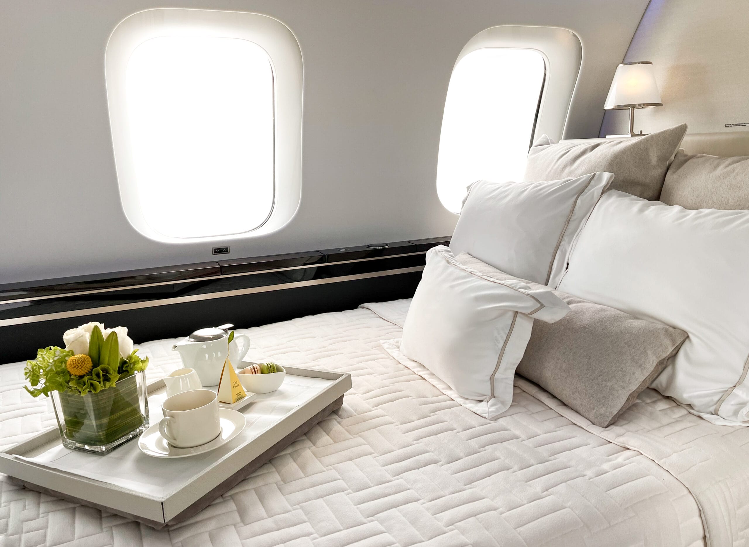 Luxurious airplane bedroom with a neatly made bed, decorative pillows, and a tray featuring tea, a vase of flowers, and pastries.