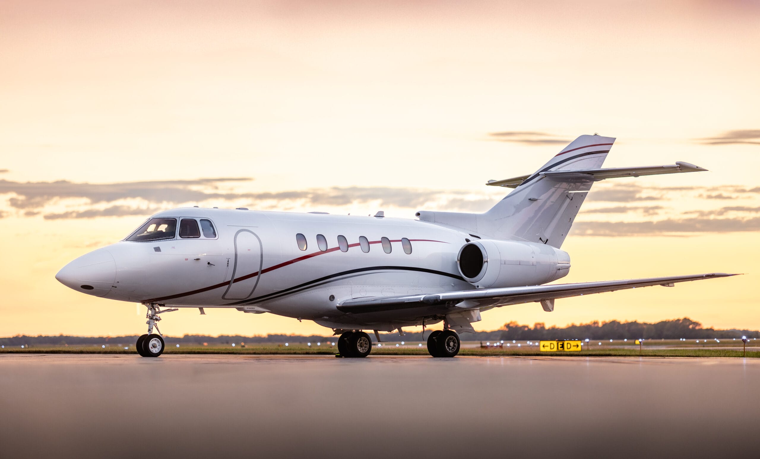 A sleek white private jet with red accents parked on a runway at sunset, showcasing a vibrant sky in the background.