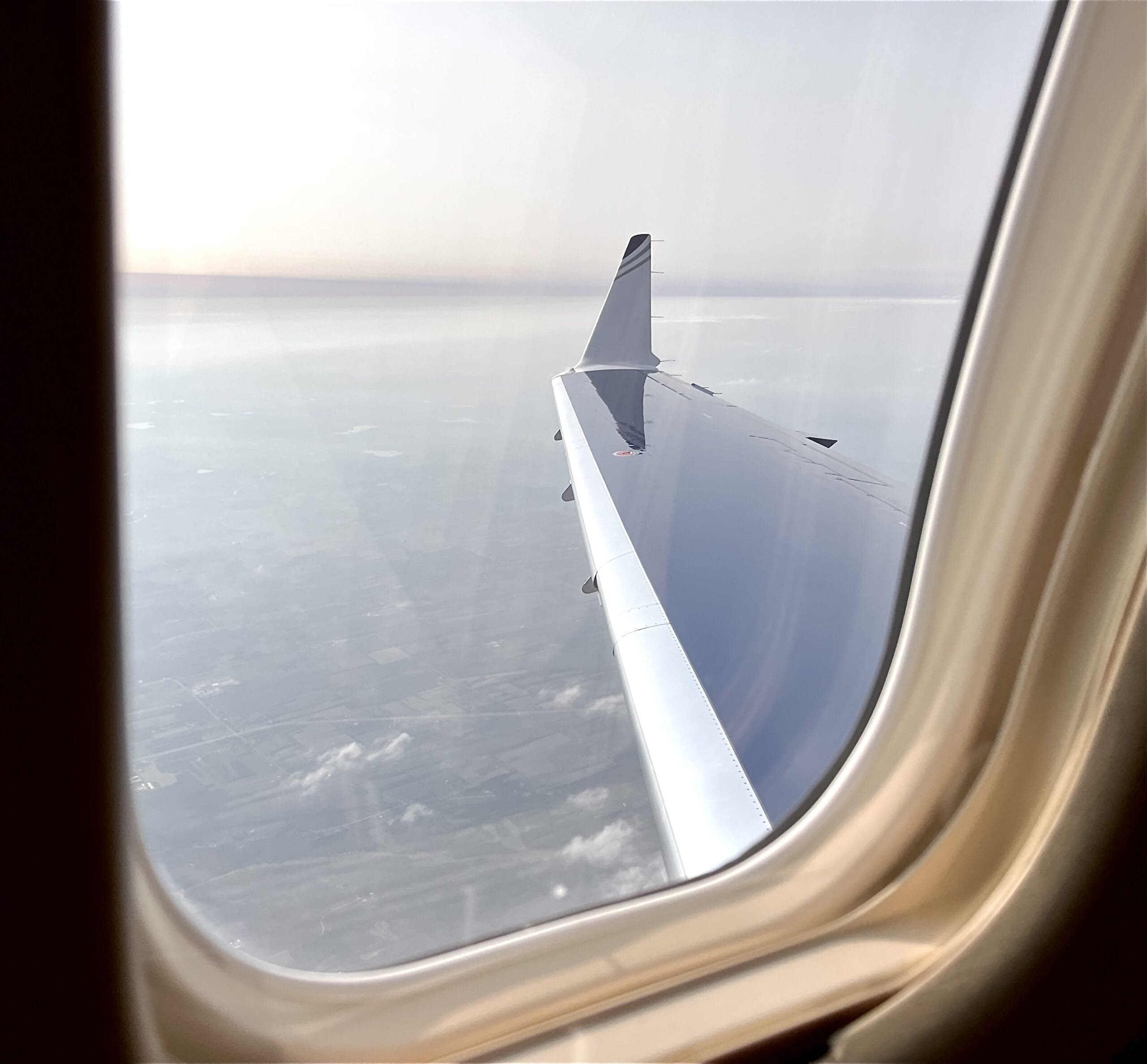 Airplane wing with a sleek design, viewed through a window, above a patchwork of fields and clouds under a hazy sky.