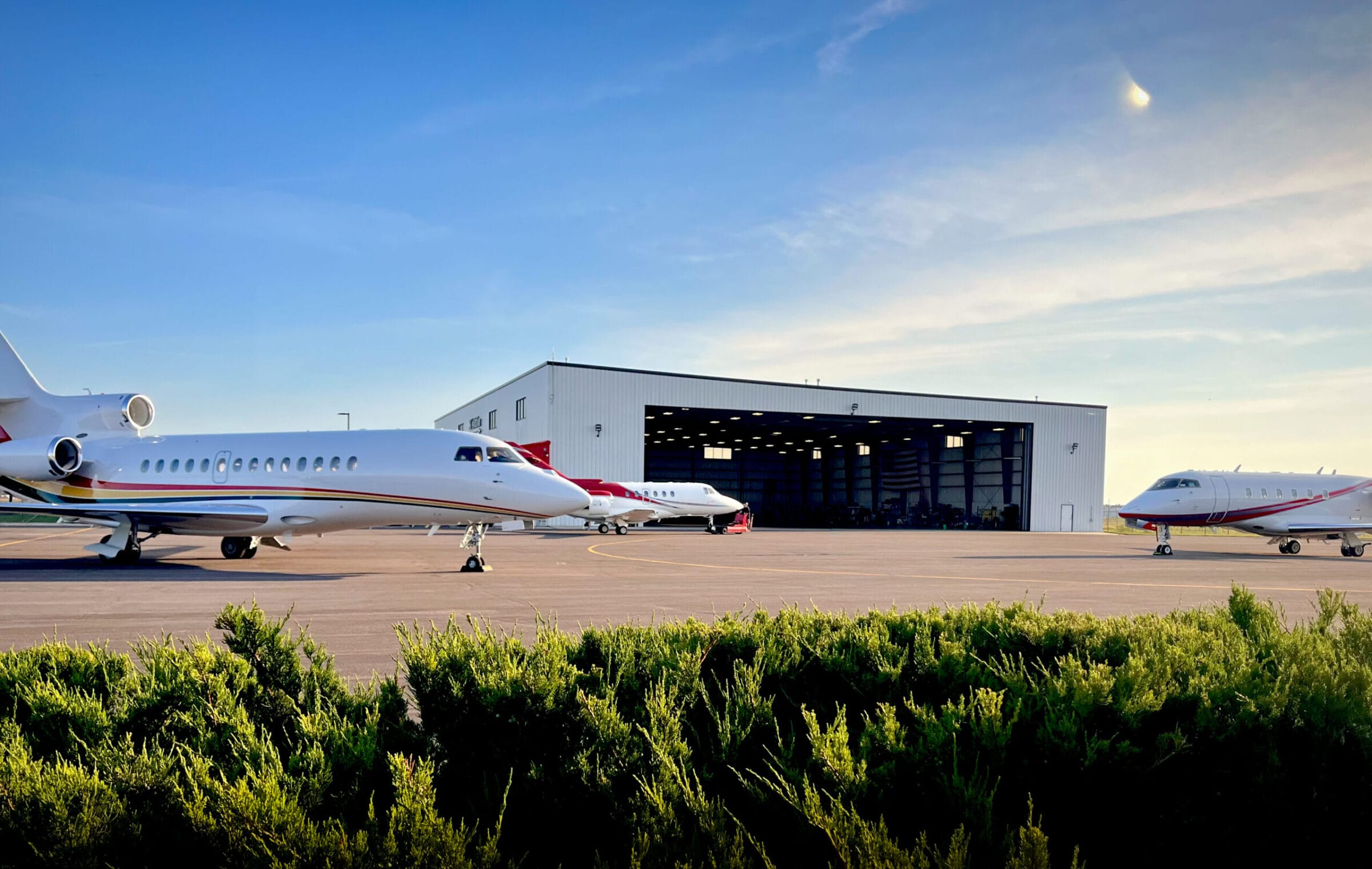 Two private jets parked on a tarmac in front of a large hangar under a clear blue sky. Green shrubs in the foreground.