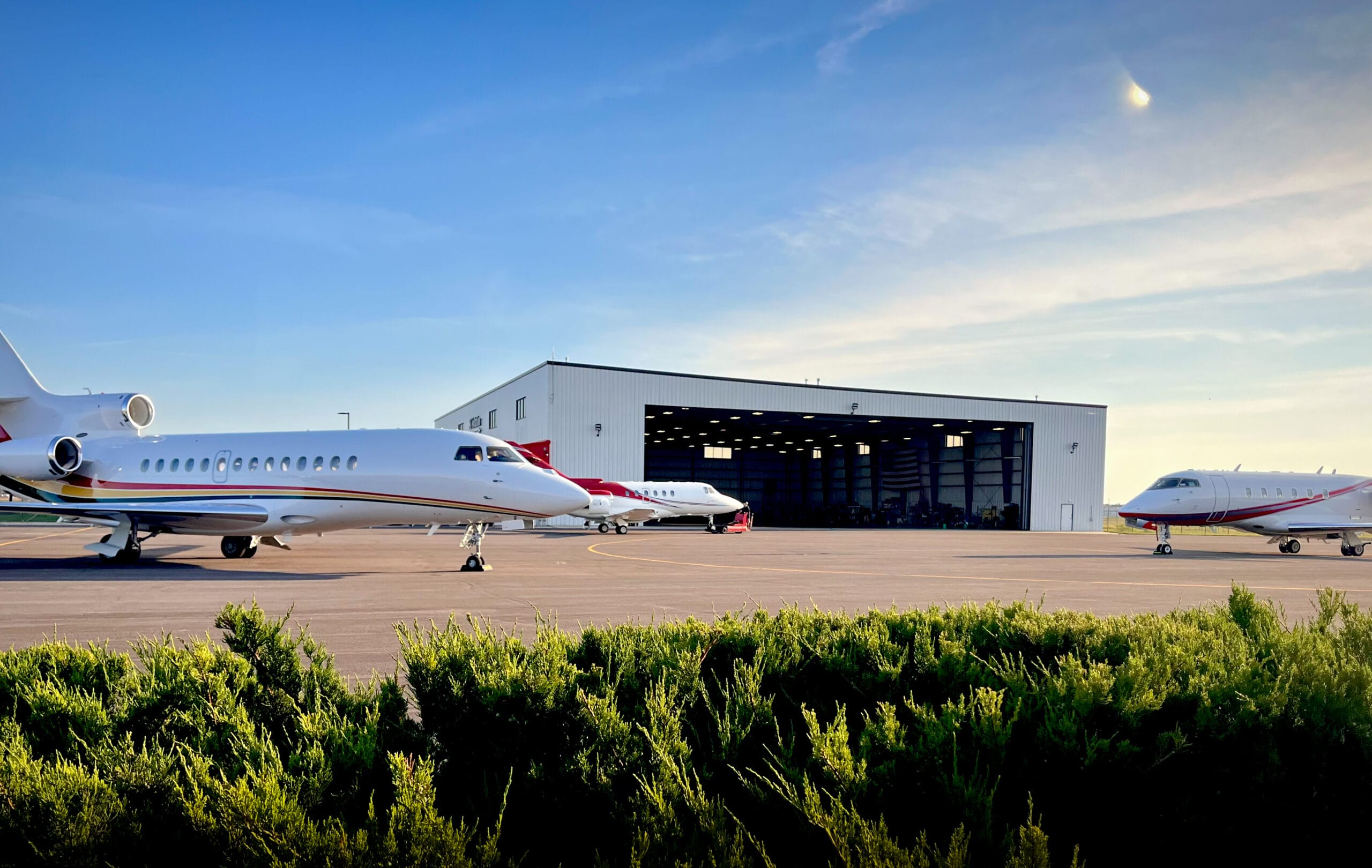Two private jets parked on a tarmac in front of a large hangar under a clear blue sky. Green shrubs in the foreground.